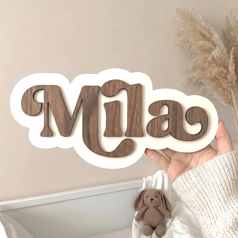 Puede incluir: Un letrero con nombre personalizado que presenta el nombre Mila en letras de madera de color marr&oacute;n oscuro montadas sobre un fondo blanco con forma de nube. Las letras tienen una fuente audaz de estilo retro con acabado liso.