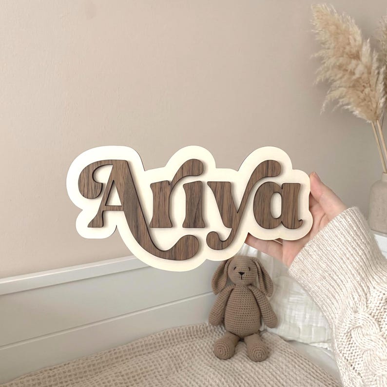 Puede incluir: Un letrero de madera personalizado con el nombre Ariya en letras de madera de color marr&oacute;n oscuro sobre un fondo de color crema. El letrero tiene una forma suave, redondeada y similar a una nube, sostenido frente a una pared lisa.
