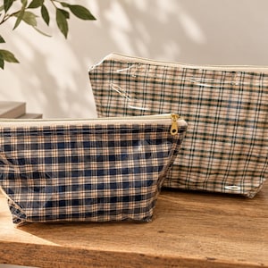 Op de afbeelding: Twee etuis met rits en een glanzende, transparante afwerking over een ruitpatroon. Het kleinere etui heeft een marineblauw en wit ruitpatroon, terwijl het grotere etui een groen, beige en wit ruitpatroon heeft. Beide hebben goudkleurige ritsen.