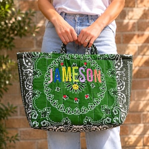 May include: A quilted tote bag made from green and black bandana fabric with a white paisley pattern. The center features the name JAMESON embroidered in multicolored letters above a small yellow smiley face and red heart accents.