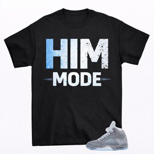 May include: Black t-shirt with the text "HIM MODE" in white and blue. A gray and light blue sneaker is shown in the lower right corner. The text has a splatter effect.