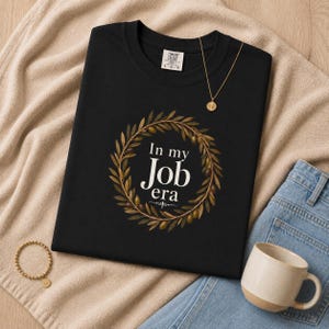 Op de afbeelding: Zwart T-shirt met de tekst "In my Job era" in een krans van olijftakken. Een gouden ketting en armband zijn zichtbaar. Een beige en bruine mok en een blauwe spijkerbroek zijn ook in de afbeelding.