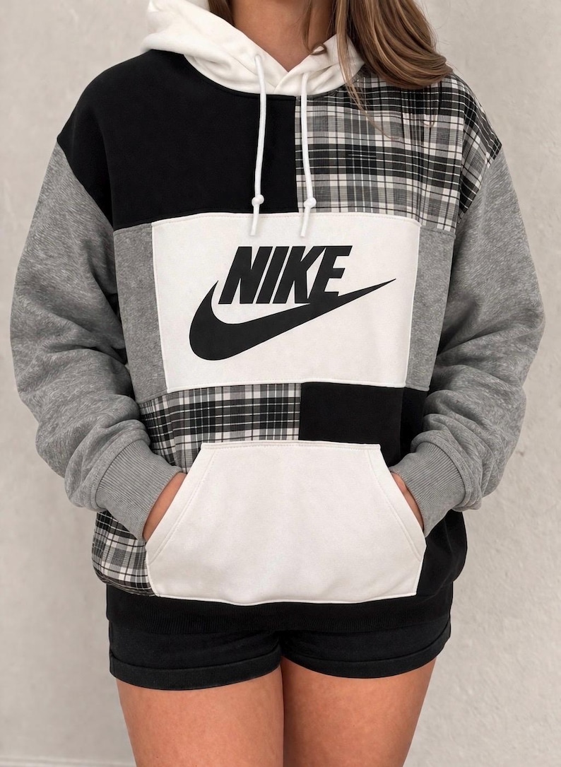 Pu&ograve; includere: Una felpa con cappuccio patchwork con cappuccio e tasca bianchi, con pannelli neri, grigi e a quadri. Il logo Nike &egrave; in nero su sfondo bianco. La felpa ha un cordino e polsini a coste.