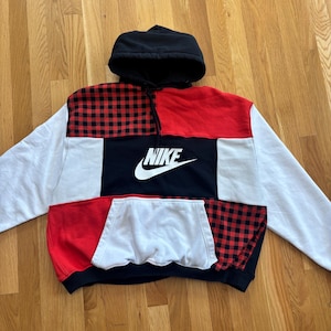 Op de afbeelding: Een patchwork hoodie met een zwarte capuchon, witte mouwen en een voorpaneel met rood en zwart buffelruit, rode, zwarte en witte secties. Het Nike-logo staat prominent in het wit op een zwart paneel.
