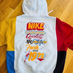 Op de afbeelding: Witte hoodie met een kleurrijk ontwerp. De achterkant heeft het Nike-logo in rood en geel, samen met de woorden "Sportswear, Quality, and Style, Fresh since 1972." De mouwen zijn geel, rood, blauw en zwart.