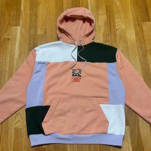 Op de afbeelding: Een colorblock hoodie in tinten perzik, wit, zwart en lavendel. De hoodie heeft een kangoeroezak en een capuchon met trekkoord. Een kleine afbeelding van een zon en regenboog is op de voorkant gedrukt.