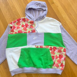 Op de afbeelding: Een patchwork hoodie in lavendel, wit en groen met bloemen- en effen kleurblokken. De hoodie heeft een kangoeroezak en een capuchon. Het Nike-logo is zichtbaar op de borst.