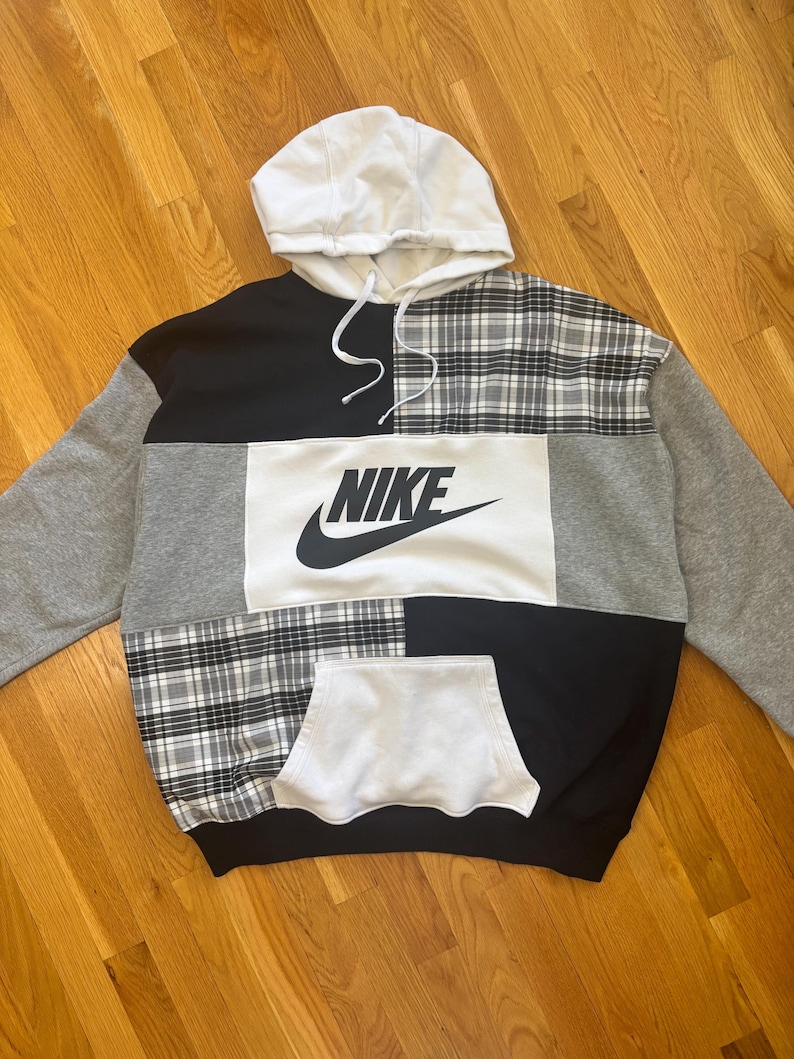 Pu&ograve; includere: Una felpa con cappuccio patchwork con cappuccio bianco e un ampio pannello bianco con il logo Nike. La felpa combina motivi neri, grigi e a quadri. Ha una tasca frontale e un cordino.