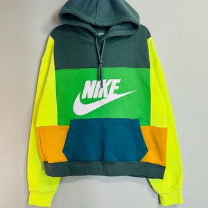 May include: A color-blocked hoodie with a green hood, green upper chest, and a green, orange, and teal lower section. The Nike logo is prominently displayed in white on the chest. The sleeves are bright yellow.