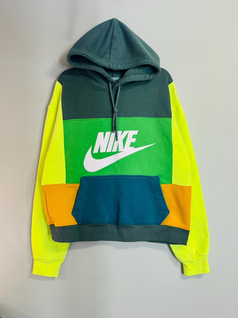 May include: A color-blocked hoodie with a green hood, green upper chest, and a green, orange, and teal lower section. The Nike logo is prominently displayed in white on the chest. The sleeves are bright yellow.