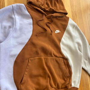 Op de afbeelding: Een colorblock hoodie in tinten bruin, gebroken wit en lavendel. De hoodie heeft een capuchon met trekkoord, een kangoeroezak en het Nike-logo op de borst. Het ontwerp is een moderne interpretatie van een klassiek sportkledingstuk.