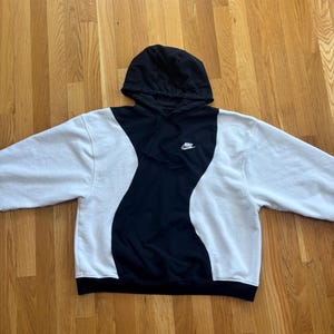 May include: A black and white Nike hoodie with a black hood and a small Nike logo on the chest. The hoodie has a unique design with a black wavy pattern down the center and white sleeves.