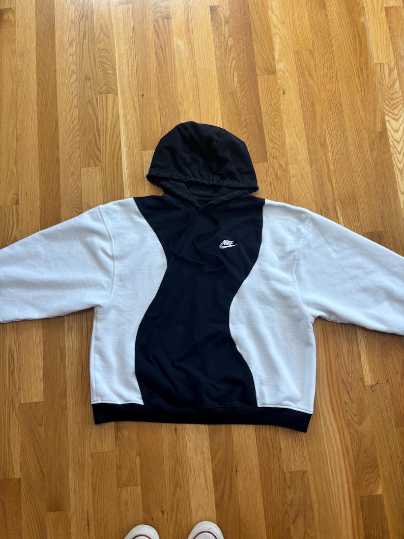 May include: A black and white Nike hoodie with a black hood and a small Nike logo on the chest. The hoodie has a unique design with a black wavy pattern down the center and white sleeves.