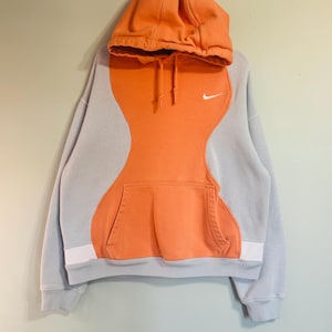 May include: A grey and orange Nike hoodie with a kangaroo pocket. The hoodie has a colour-block design with orange in the centre and grey on the sleeves and hood. A small white Nike swoosh logo is on the chest.