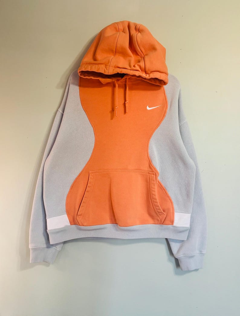 May include: A grey and orange Nike hoodie with a kangaroo pocket. The hoodie has a colour-block design with orange in the centre and grey on the sleeves and hood. A small white Nike swoosh logo is on the chest.
