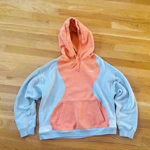 May include: A light blue and coral-coloured Nike hoodie with a front pocket and a drawstring hood. The hoodie features a white Nike logo on the chest. The design has a unique colour-block pattern.