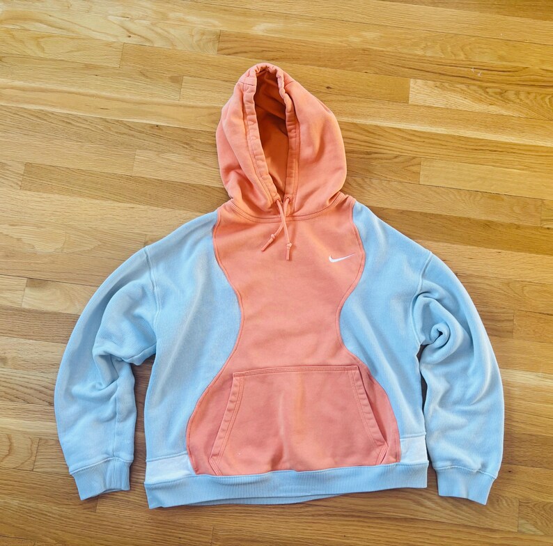 May include: A light blue and coral-coloured Nike hoodie with a front pocket and a drawstring hood. The hoodie features a white Nike logo on the chest. The design has a unique colour-block pattern.