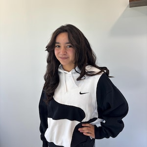 Puede incluir: Una sudadera con capucha y manga larga que presenta un diseño de bloques de color en blanco y negro. El panel frontal muestra un patrón curvo en forma de onda que separa los dos colores, con un pequeño logotipo Nike negro bordado en el pecho.