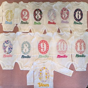 Może przedstawiać: Kolekcja białych body dla niemowląt z długimi i kr&oacute;tkimi rękawami, z nadrukowaną kwiatowym wzorem liczbą od 1 do 11, reprezentującą miesiące i rok. Liczby są w r&oacute;żnych kolorach, ze słowem "Months" lub "Year" poniżej.