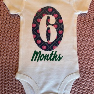 Może przedstawiać: Białe body dla dziecka z dużym owalnym wzorem z białym numerem "6", otoczonym wzorem czerwonych jabłek na ciemnoniebieskim tle. Słowo "Months" jest wydrukowane na zielono poniżej owalu.