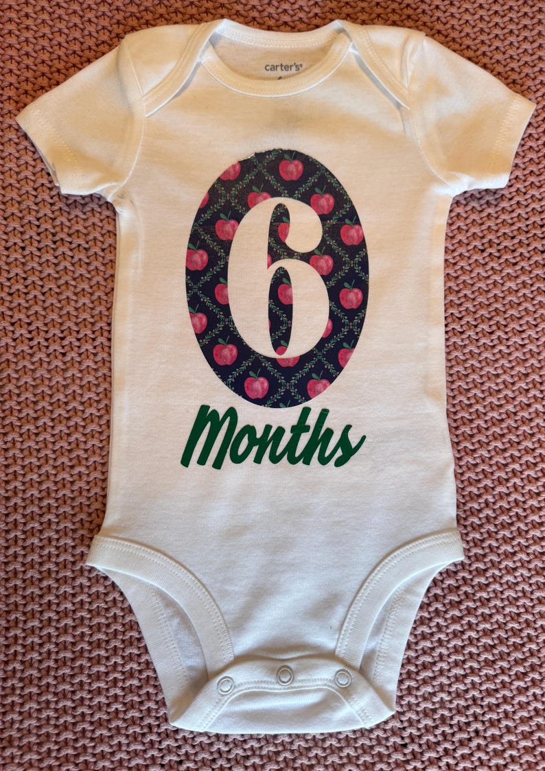 Może przedstawiać: Białe body dla dziecka z dużym owalnym wzorem z białym numerem "6", otoczonym wzorem czerwonych jabłek na ciemnoniebieskim tle. Słowo "Months" jest wydrukowane na zielono poniżej owalu.