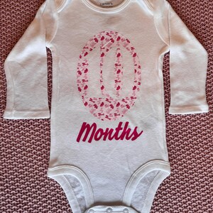 Może przedstawiać: Białe body dla dziecka z długim rękawem, z r&oacute;żowym kwiatowym wzorem tworzącym cyfrę 10. Poniżej napis &bdquo;Months&rdquo; w r&oacute;żowym skrypcie. Body wykonane jest z miękkiego materiału, dla dziesięciomiesięcznego dziecka.