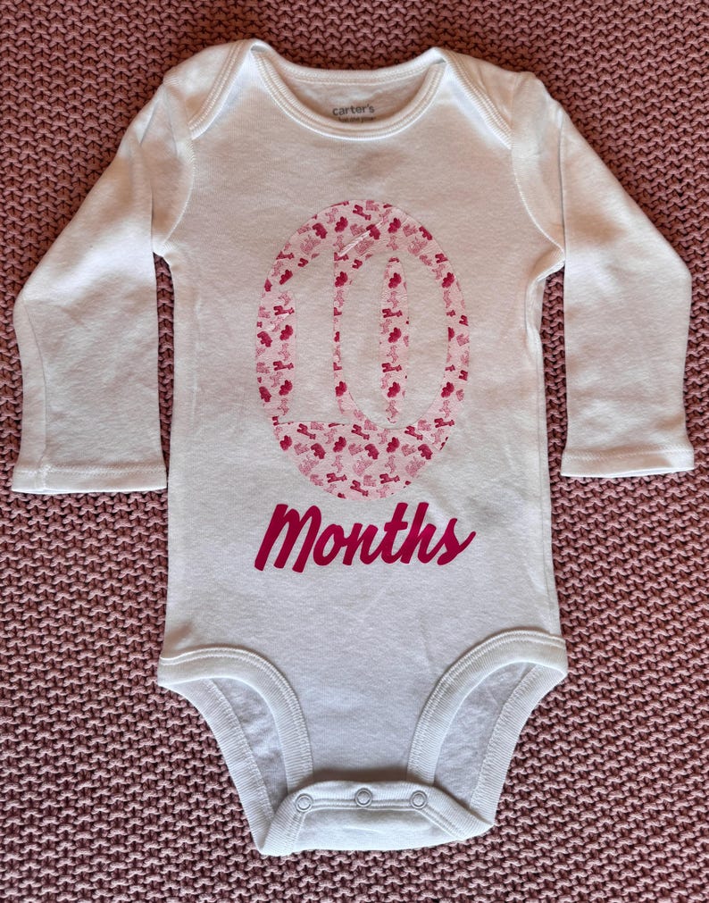 Może przedstawiać: Białe body dla dziecka z długim rękawem, z r&oacute;żowym kwiatowym wzorem tworzącym cyfrę 10. Poniżej napis &bdquo;Months&rdquo; w r&oacute;żowym skrypcie. Body wykonane jest z miękkiego materiału, dla dziesięciomiesięcznego dziecka.
