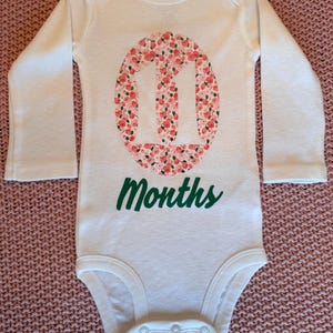 Może przedstawiać: Białe body dla dziecka z długim rękawem z zielonym napisem "11 Months". Liczba "11" znajduje się w owalu wypełnionym r&oacute;żowo-czerwonym wzorem kwiatowym. Body posiada zapięcie na zatrzaski.