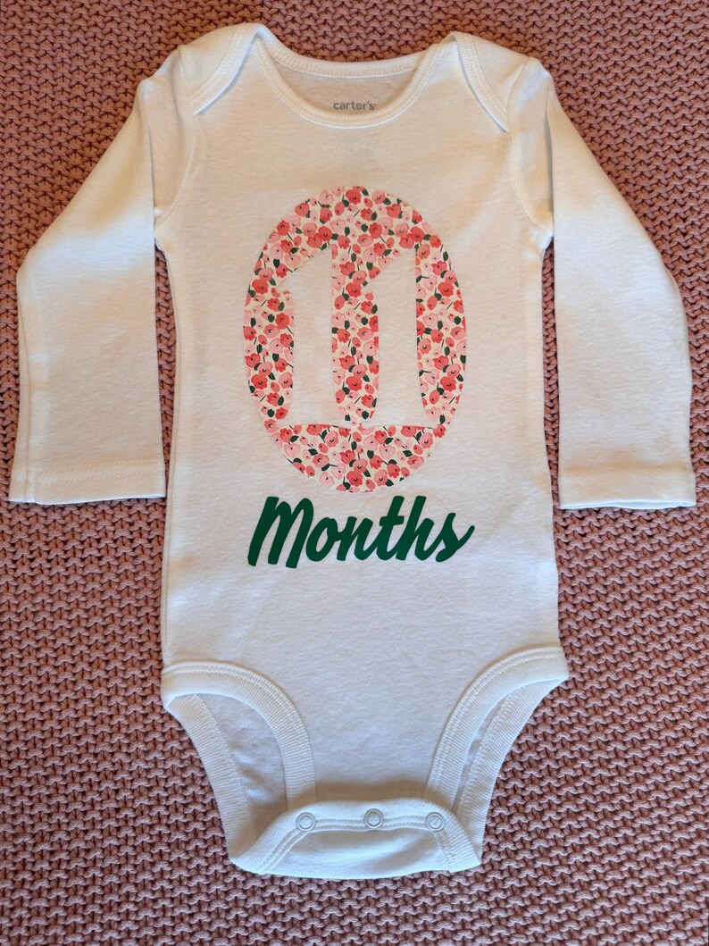 Może przedstawiać: Białe body dla dziecka z długim rękawem z zielonym napisem "11 Months". Liczba "11" znajduje się w owalu wypełnionym r&oacute;żowo-czerwonym wzorem kwiatowym. Body posiada zapięcie na zatrzaski.