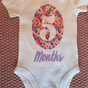 Może przedstawiać: Białe body dla dziecka z kwiatowym wzorem "5" na środku. Numer wypełniony jest r&oacute;żowymi, czerwonymi, fioletowymi i ż&oacute;łtymi kwiatami. Słowo "Months" jest wydrukowane poniżej w lawendowym skrypcie.