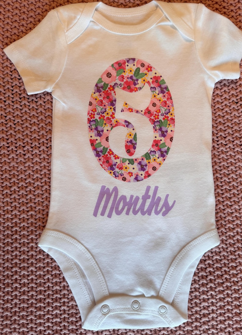 Może przedstawiać: Białe body dla dziecka z kwiatowym wzorem "5" na środku. Numer wypełniony jest r&oacute;żowymi, czerwonymi, fioletowymi i ż&oacute;łtymi kwiatami. Słowo "Months" jest wydrukowane poniżej w lawendowym skrypcie.