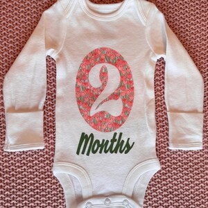 Może przedstawiać: Białe body dla dziecka z długim rękawem. Numer "2" jest wyświetlany we wzorze owalu z kwiatami. Słowo "Months" jest wydrukowane na zielono poniżej. Body ma zapięcia na zatrzaski na dole.