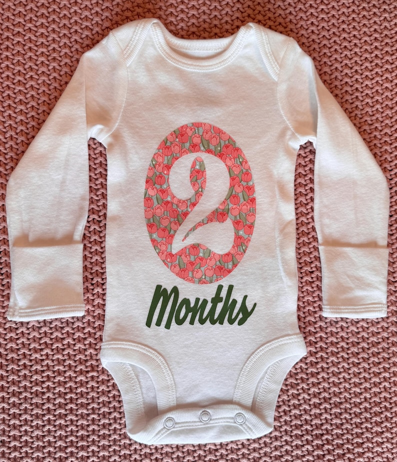 Może przedstawiać: Białe body dla dziecka z długim rękawem. Numer "2" jest wyświetlany we wzorze owalu z kwiatami. Słowo "Months" jest wydrukowane na zielono poniżej. Body ma zapięcia na zatrzaski na dole.