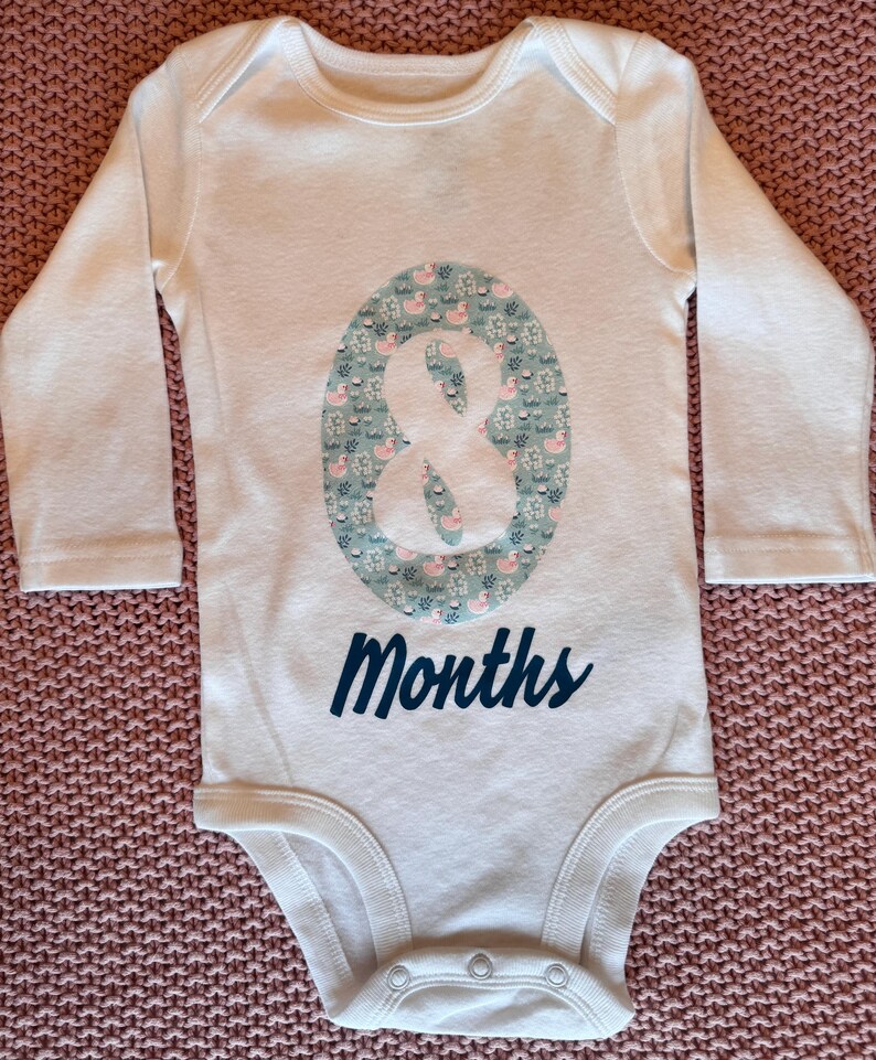 Może przedstawiać: Białe body dla dziecka z długimi rękawami. Posiada duży owalny wz&oacute;r z motywem kaczek i element&oacute;w kwiatowych w odcieniach niebieskiego i r&oacute;żowego. Słowo "Months" jest wydrukowane w kolorze granatowym.