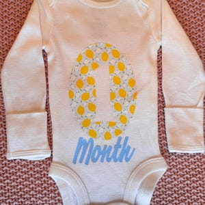 Może przedstawiać: Białe body dla dziecka z długimi rękawami. Body posiada dużą cyfrę "1" wypełnioną ż&oacute;łtymi cytrynami i jasnoniebieskim tłem. Słowo "Month" jest napisane jasnoniebieską czcionką poniżej cyfry.