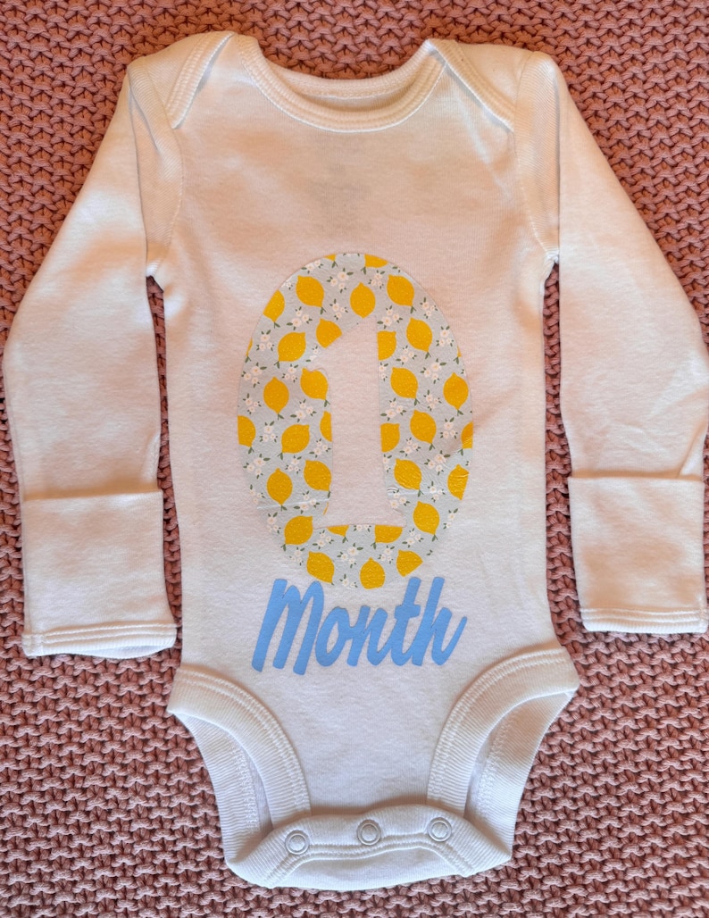 Może przedstawiać: Białe body dla dziecka z długimi rękawami. Body posiada dużą cyfrę "1" wypełnioną ż&oacute;łtymi cytrynami i jasnoniebieskim tłem. Słowo "Month" jest napisane jasnoniebieską czcionką poniżej cyfry.