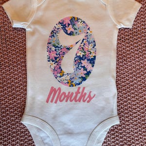 Może przedstawiać: Białe body dla dziecka z kwiatowym wzorem "7" w kolorze niebieskim, r&oacute;żowym i ż&oacute;łtym. Słowo "Months" jest wydrukowane na r&oacute;żowo pod numerem. Body ma kr&oacute;tkie rękawy i zatrzaski na dole.