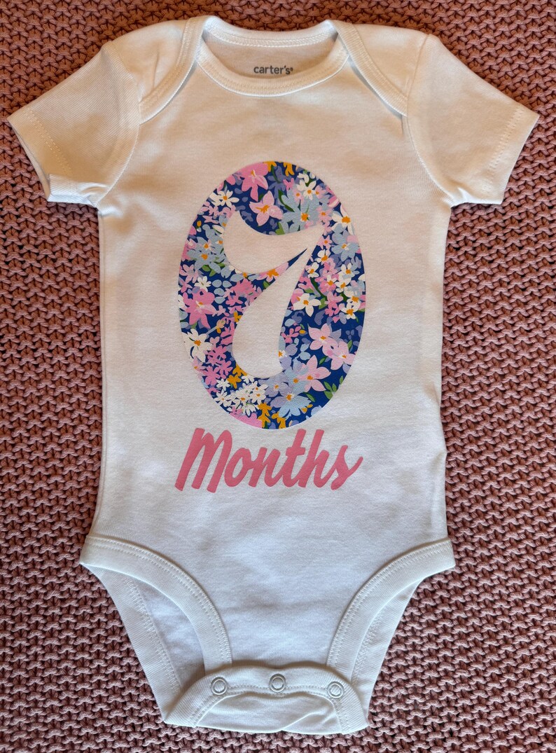 Może przedstawiać: Białe body dla dziecka z kwiatowym wzorem "7" w kolorze niebieskim, r&oacute;żowym i ż&oacute;łtym. Słowo "Months" jest wydrukowane na r&oacute;żowo pod numerem. Body ma kr&oacute;tkie rękawy i zatrzaski na dole.