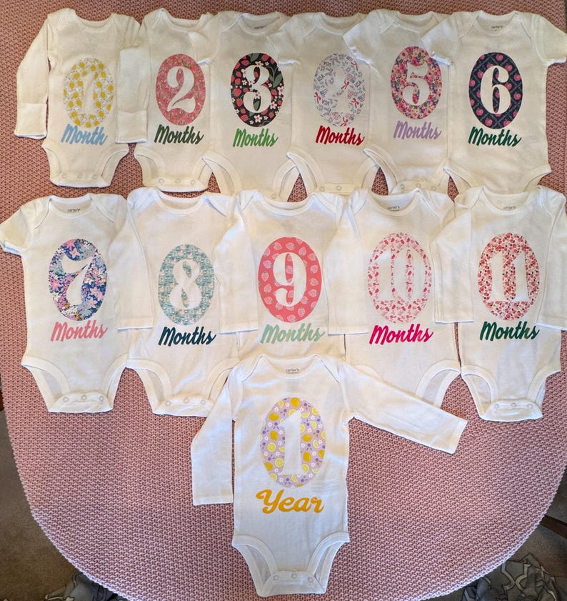 Może przedstawiać: Kolekcja białych body dla niemowląt z długimi i kr&oacute;tkimi rękawami, z nadrukowaną kwiatowym wzorem liczbą od 1 do 11, reprezentującą miesiące i rok. Liczby są w r&oacute;żnych kolorach, ze słowem "Months" lub "Year" poniżej.