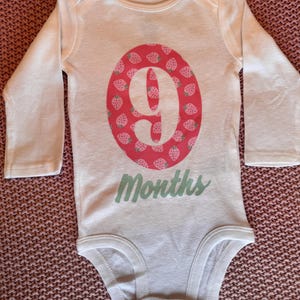 Może przedstawiać: Białe body dla dziecka z długim rękawem. Posiada r&oacute;żowy owal ze wzorem truskawek i cyfrą 9. Słowo "Months" jest napisane na dole jasną zielenią.