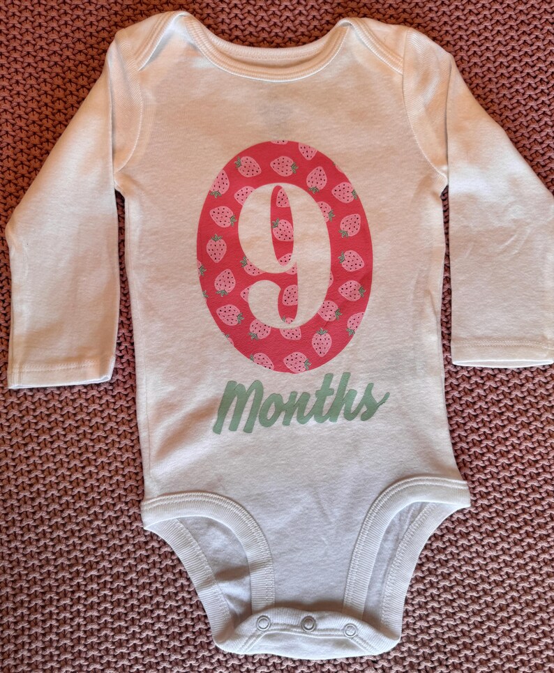 Może przedstawiać: Białe body dla dziecka z długim rękawem. Posiada r&oacute;żowy owal ze wzorem truskawek i cyfrą 9. Słowo "Months" jest napisane na dole jasną zielenią.