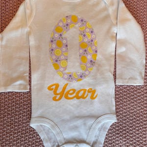 Może przedstawiać: Białe body niemowlęce z długim rękawem z dużym numerem "1" pośrodku, ozdobione cytrynami i fioletowymi kwiatami. Słowo "Year" jest wydrukowane na pomarańczowo poniżej. Body jest wykonane z miękkiego materiału.