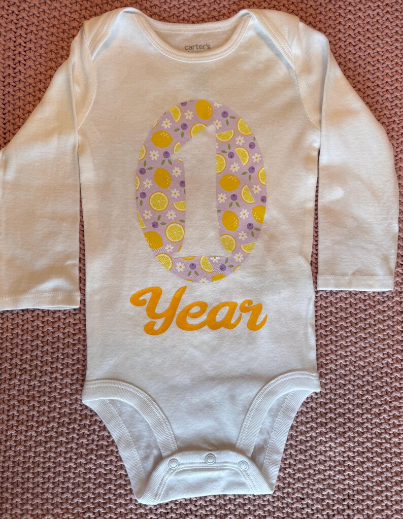 Może przedstawiać: Białe body niemowlęce z długim rękawem z dużym numerem "1" pośrodku, ozdobione cytrynami i fioletowymi kwiatami. Słowo "Year" jest wydrukowane na pomarańczowo poniżej. Body jest wykonane z miękkiego materiału.