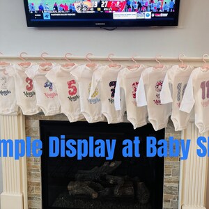 Może przedstawiać: Białe body dla niemowląt z nadrukowanymi cyframi i słowem "Months", prezentowane na r&oacute;żowych wieszakach. Widoczne są cyfry 1, 3, 4, 5, 7 i 11. Widoczny jest r&oacute;wnież tekst "Example Display at Baby Show".