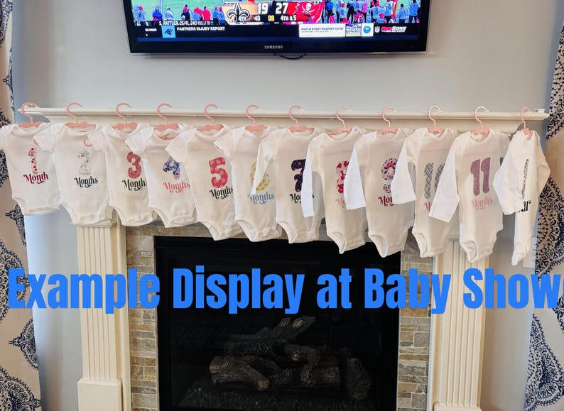 Może przedstawiać: Białe body dla niemowląt z nadrukowanymi cyframi i słowem "Months", prezentowane na r&oacute;żowych wieszakach. Widoczne są cyfry 1, 3, 4, 5, 7 i 11. Widoczny jest r&oacute;wnież tekst "Example Display at Baby Show".