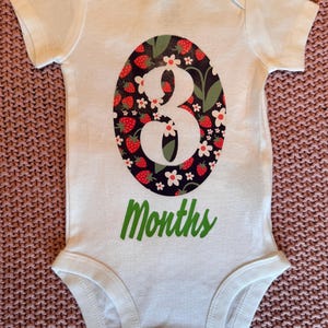 Może przedstawiać: Białe body dla dziecka z dużym owalnym wzorem z białym numerem "3", otoczonym truskawkami i kwiatami na ciemnym tle. Słowo "Months" jest wydrukowane na zielono poniżej.