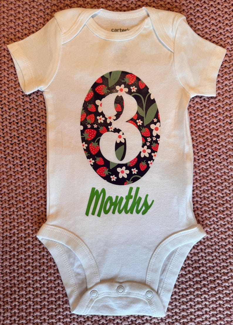 Może przedstawiać: Białe body dla dziecka z dużym owalnym wzorem z białym numerem "3", otoczonym truskawkami i kwiatami na ciemnym tle. Słowo "Months" jest wydrukowane na zielono poniżej.