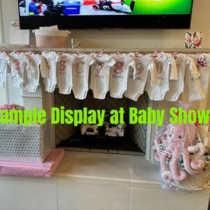 Może przedstawiać: Ekspozycja na baby shower z białymi body z nadrukowanymi cyframi i miesiącami, wiszącymi na drążku. Kosz prezentowy z r&oacute;żową pluszową ośmiornicą i innymi przedmiotami znajduje się po prawej stronie. Pudełko prezentowe znajduje się po lewej stronie.