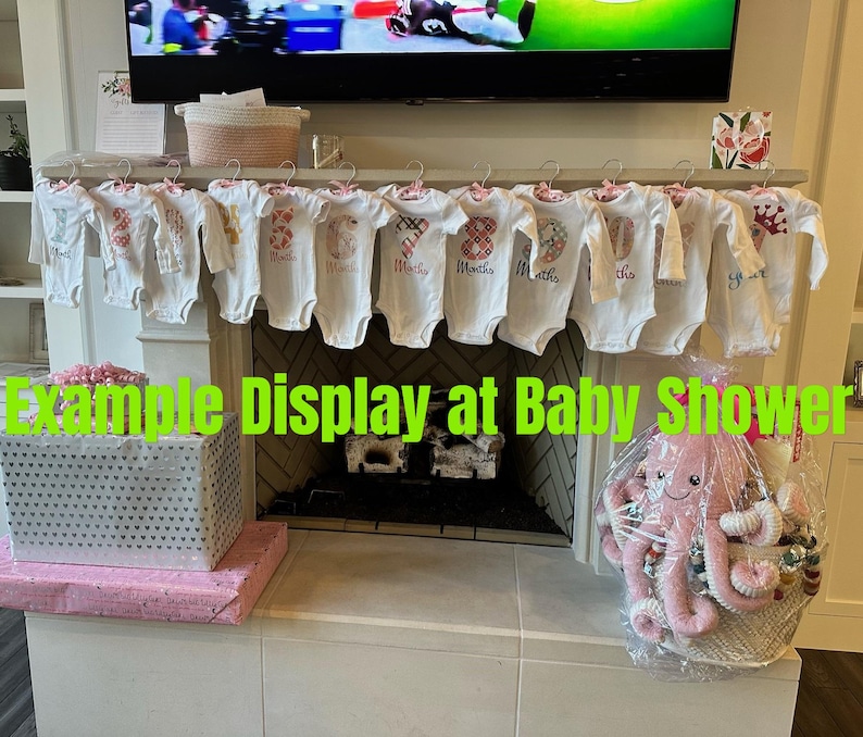 Może przedstawiać: Ekspozycja na baby shower z białymi body z nadrukowanymi cyframi i miesiącami, wiszącymi na drążku. Kosz prezentowy z r&oacute;żową pluszową ośmiornicą i innymi przedmiotami znajduje się po prawej stronie. Pudełko prezentowe znajduje się po lewej stronie.