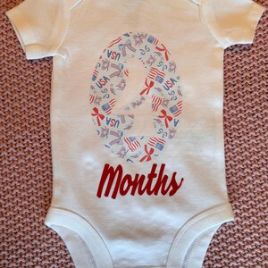 Może przedstawiać: Białe body dla dziecka z patriotycznym wzorem. Numer "2" wypełniony jest grafiką w kolorach czerwonym, białym i niebieskim w stylu USA. Słowo "Months" jest napisane czerwoną czcionką poniżej. Ubranko dla dziecka.